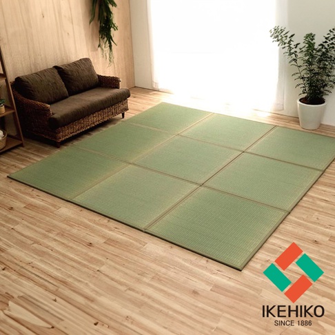 一張綠色藺草組合式三折疊榻榻米地墊，鋪設在淺木紋地板上，一旁有深棕色藤編沙發和盆栽植物。