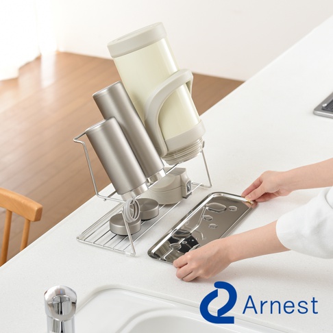 Arnest 牌不鏽鋼雙層瀝水架，展示架上有兩個銀色和一個米色保溫瓶，以及一個裝有水的銀色托盤，由一名女子用手調整。