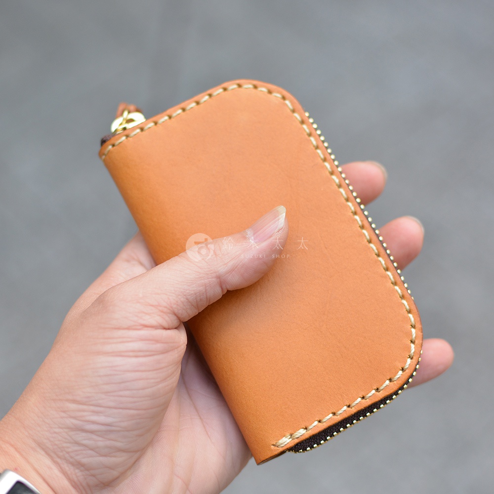 手持一個駝色皮革鑰匙包，包體呈現橢圓形，邊緣有精緻的縫線裝飾，拉鍊為金色。