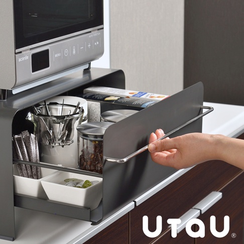 灰色金屬材質的抽屜式收納櫃，內有兩個白色小方碗，裝有茶包或咖啡包，以及透明玻璃罐裝有咖啡豆，上方置有咖啡濾網架，櫃體上方放置著微波爐。