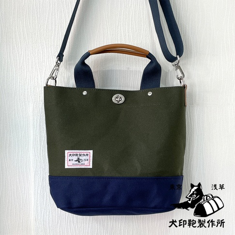 犬印純棉帆布兩用雙色托特包，主體為軍綠色帆布，底部拼接海軍藍色帆布，配有海軍藍色織帶提把和肩帶，以及棕色皮革提把。包身中央有金屬旋轉鎖扣，左側有一個印有品牌標誌的白色布標。