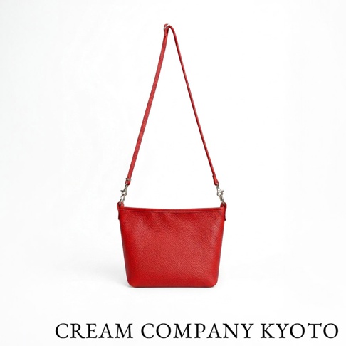 這是一款由日本 CREAM COMPANY KYOTO 出品的紅色皮革隨身側背包，包身採用質感細緻的粒面牛皮製成，呈現簡約俐落的梯形輪廓。包包配備可拆卸的同色系皮革肩背帶，兩側以金屬扣環連接，整體設計輕便實用，底部印有品牌名稱。
