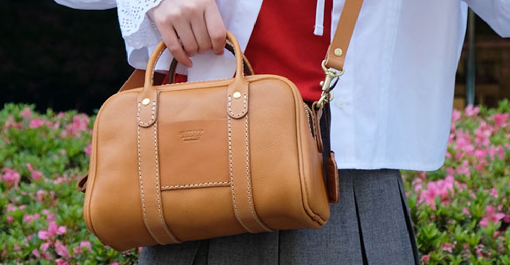 一名女子提著一個HERZ品牌的咖啡色皮革迷你波士頓包，包包上有明顯的縫線和壓印Logo，背景是綠色和粉色花叢。