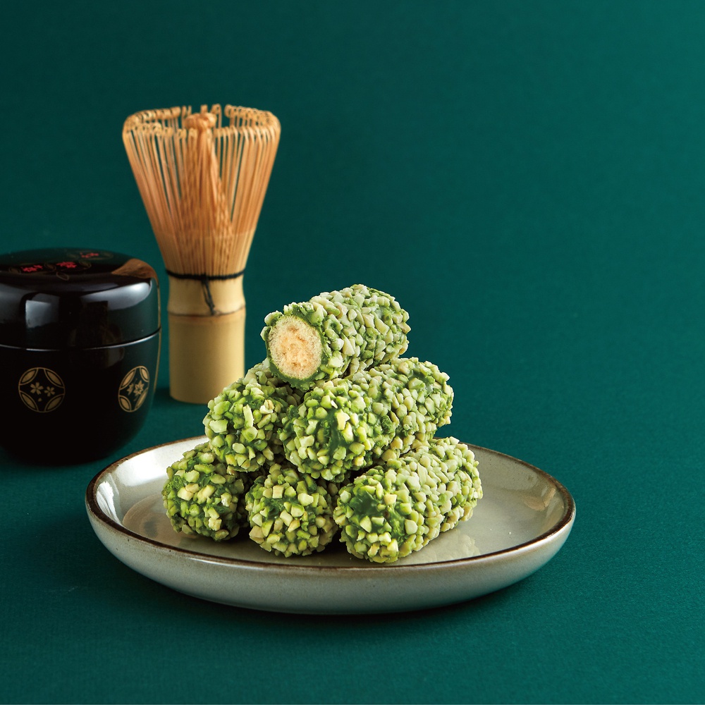 堆疊的抹茶杏仁卷，外層裹滿翠綠色碎杏仁，內餡為淺黃色，旁邊擺放黑色抹茶罐和竹製茶筅。