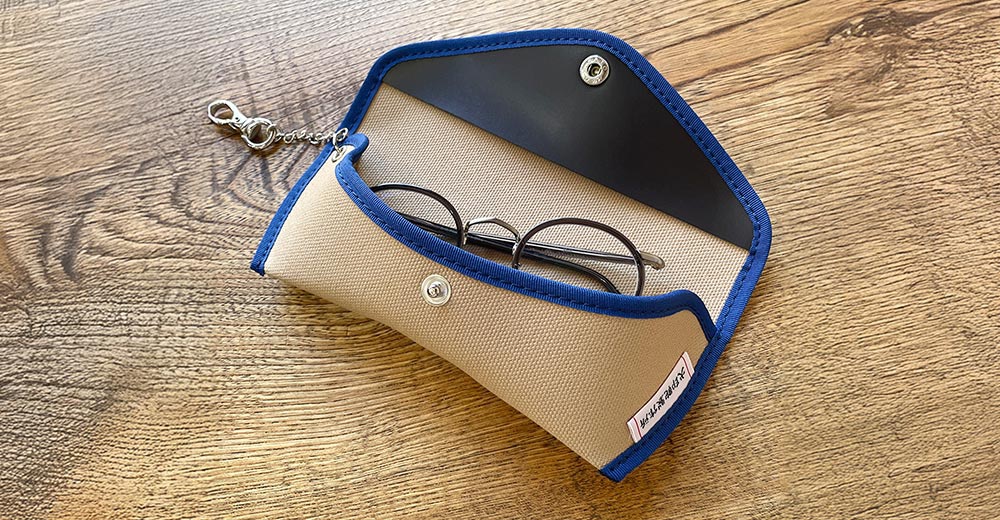 純棉帆布製成的卡其色眼鏡收納盒，飾有藍色滾邊，盒蓋內有黑色皮革襯裡，並有銀色按扣，掛繩上帶有鑰匙圈，盒蓋一角印有犬印純綿6號帆布的日文標誌。