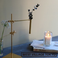 黃銅材質的支架上，夾著一個細長的黃銅花筒，花筒內插著一株黑色枝葉植物。背景為淺灰色牆面，桌面上有一玻璃瓶裝白色小花、一本雜誌和一罐點燃的蠟燭。