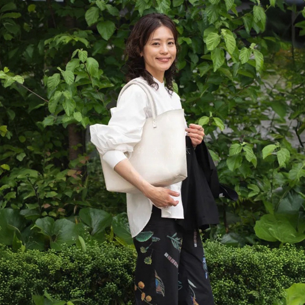 一位女士手持淺色托特包，包款為簡約的肩背手提設計，搭配白色寬袖上衣與印花長褲，背景為綠意盎然的植物。