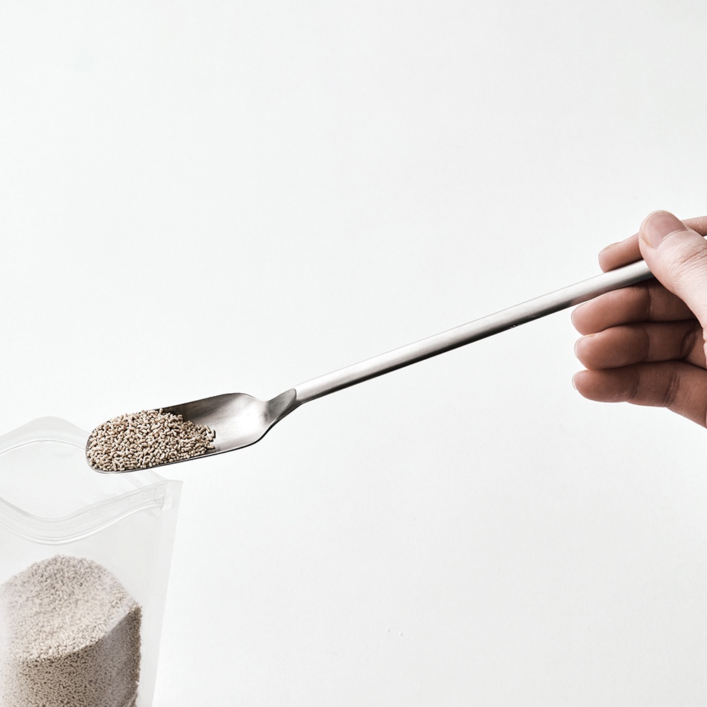 一只手拿著一支日製不鏽鋼小長勺，勺內盛有細小顆粒狀的淺褐色物質，勺口邊緣略微彎曲，勺柄細長，與白色背景形成對比。