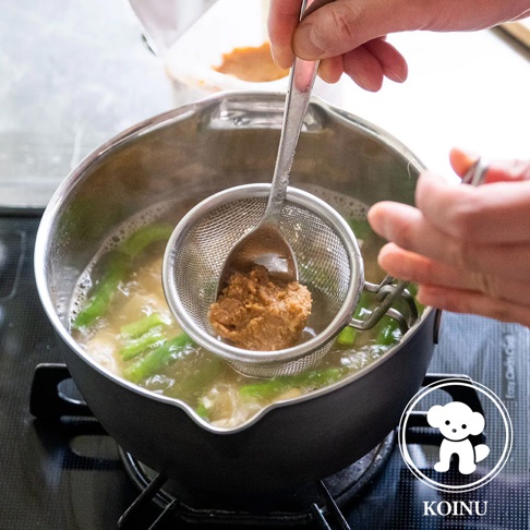 不鏽鋼雪平鍋內煮著綠色蔬菜和湯，湯面上飄著少量泡沫。一隻手正用湯匙將味噌醬透過濾網加入鍋中。