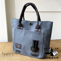 藍灰色帆布水桶托特包，配有黑色織帶提把、銀色鉚釘裝飾，正面有一個翻蓋口袋，口袋內有黑色皮革筆插，包身綴有「大印範製作所」標籤。