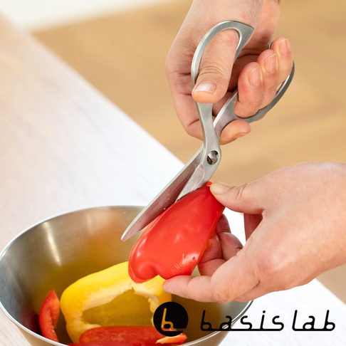 一雙銀色不鏽鋼料理剪刀，正在剪切紅黃色甜椒，剪刀的一側是食物，另一側是手，剪刀由螺絲固定，刀刃鋒利。