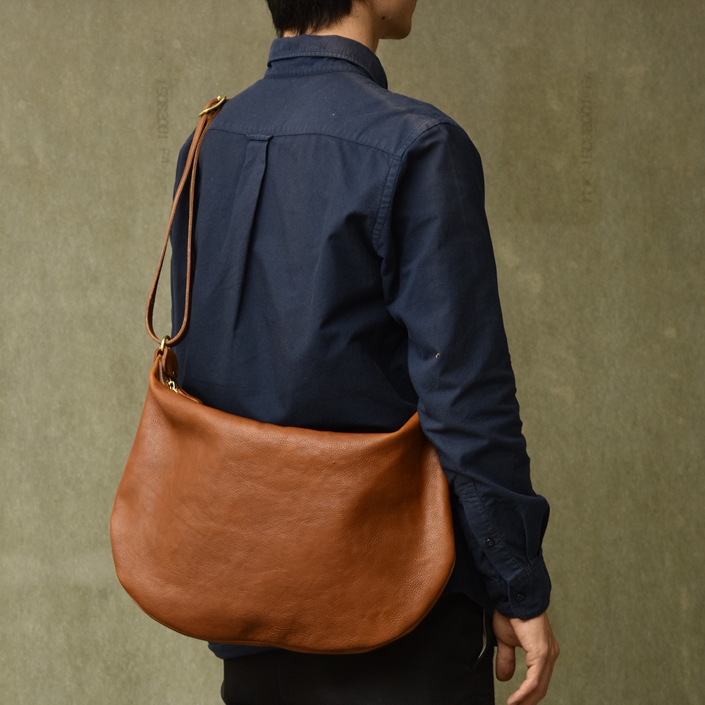 職人手作皮革Organ單肩半月包，採用柔軟的棕褐色皮革製作，呈現自然紋理，包身呈現半月形，配有可調節的皮革肩帶和金屬拉鍊。