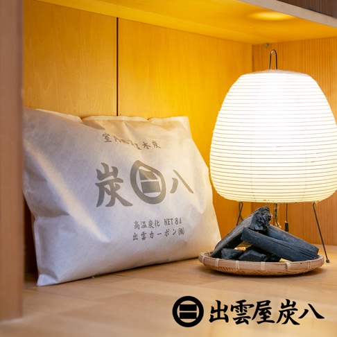 一包米白色紙袋裝的調濕木炭，袋上印有「炭八」字樣及日文說明，旁邊有一個竹篩，篩內盛放著幾塊深黑色、粗糙質地的木炭，背景為黃色木質層架與一盞白色紙罩的檯燈。
