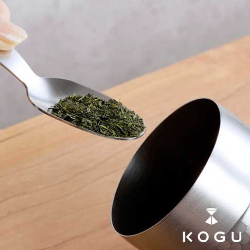日製18-8不鏽鋼錘目紋茶葉罐，附有茶匙，茶匙中裝有綠色茶葉，準備放入罐中。