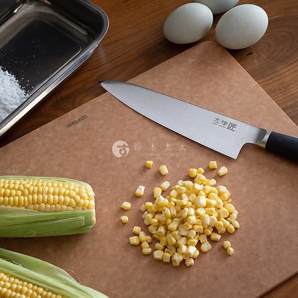 一把不鏽鋼材質的都 / Santoku 萬用刀，刀刃上有明顯的紋路，黑色握柄，刀身印有「忠 匠」字樣。刀旁是玉米粒、玉米筍和雞蛋。
