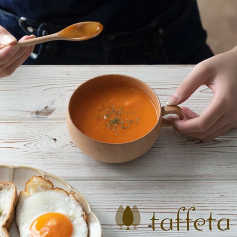 圓形楓木湯杯盛有橙紅色濃湯，湯面撒有綠色香料。湯杯配有木質手柄，由手持。背景為白色木紋桌面。