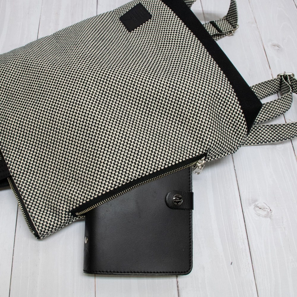 三河木綿刺子織手提後背包，布料為黑白格紋圖案，搭配黑色邊條與拉鍊。一黑色皮質筆記本置於背包前方拉鍊處。