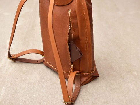 職人手作皮革後背包，採簡約褶皺風設計，主體為溫潤的棕色皮革，配有黃銅拉鍊及可調節的皮革肩帶。包側有一個拉鍊口袋，露出一個同色系的皮革零錢包。