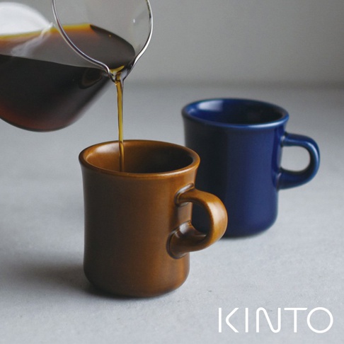 兩款 SCS 馬克杯，一款為暖棕色，另一款為深藍色，皆為陶瓷材質，造型簡潔，有圓形把手。其中暖棕色馬克杯正被傾倒咖啡。