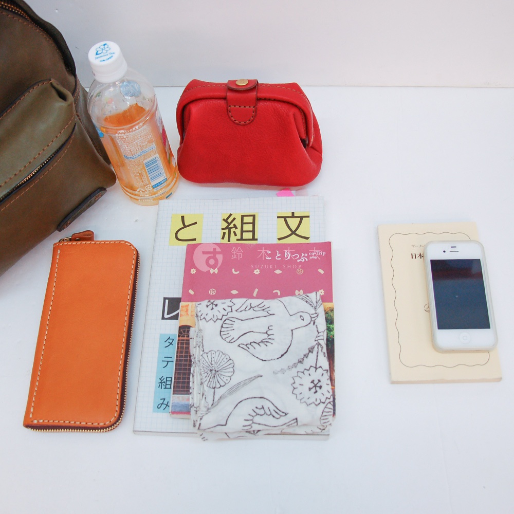 皮革後背包（棕色）、紅色皮革零錢包、橘色皮革長夾、日文雜誌、印有鳥類圖案的布織品，以及白色智慧型手機。