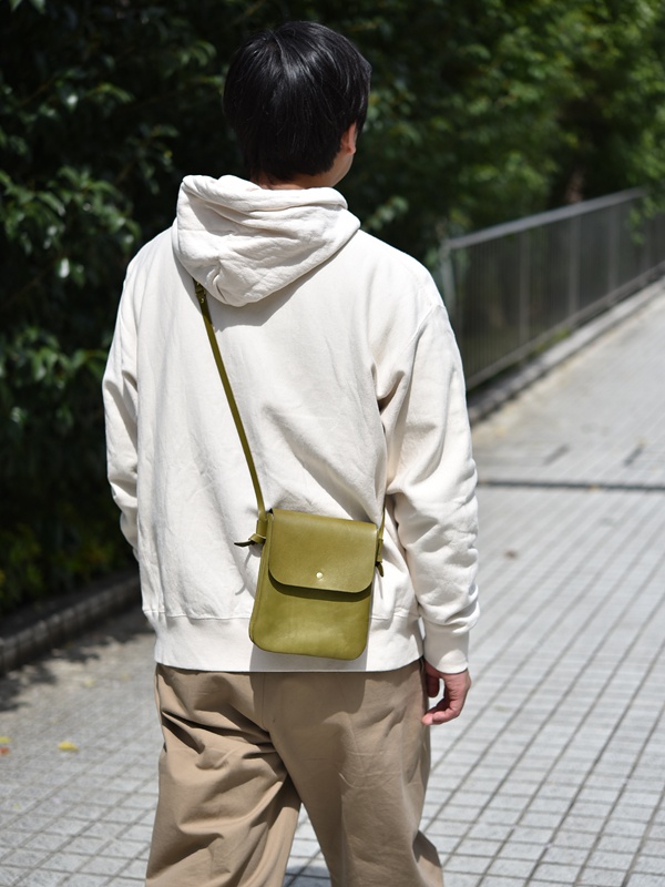 橄欖綠皮革橫式隨行袋，帶有金色按扣，斜挎於身穿米白色連帽衫的模特身上。