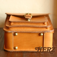 職人手作皮革單肩兩用小方包，採用鞣製皮革材質，呈現經典棕褐色，包身結構層次分明，配有金屬釦環和铆釘裝飾，整體風格簡約復古。