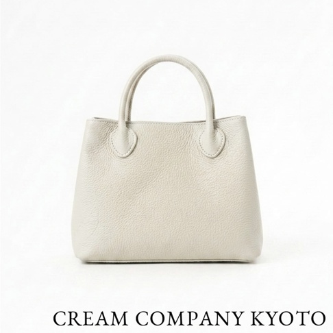 這是一張正面視角的 CREAM COMPANY KYOTO 品牌手提托特包，包身採用淺米白色荔枝紋皮革製成，設計簡約精緻，設有圓弧形手提把，下方印有黑色品牌名稱文字。