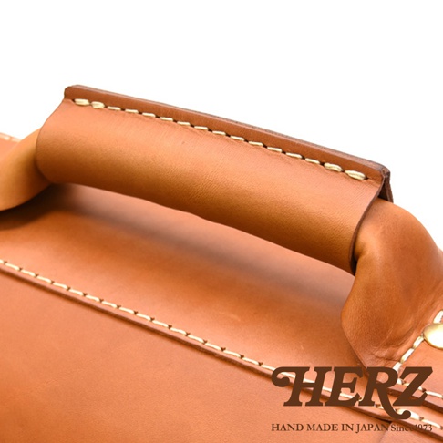 職人手作皮革經典雙扣兩用單肩包 A-2，棕色光滑皮革提把，配有米白色車線，展現細緻工藝。包體為淺棕色皮革，邊緣有對比色車縫線，整體風格經典耐看。