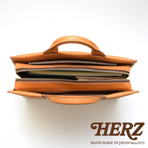 日本HERZ手工皮革簡約公事包，頂部視角，皮革顏色為棕色，包內裝有文件夾和筆記本，並有品牌HERZ和HAND MADE IN JAPAN Since1973的標誌。
