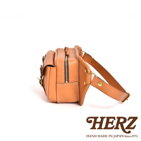 職人手作皮革騎士隨身斜背包，採用天然皮革製成，呈現溫潤的淺棕色。包款側邊帶有拉鍊開口，並以金屬釦件和皮革帶固定，設計簡潔實用。