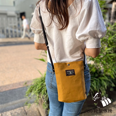 一個女人身穿白色泡泡袖上衣和牛仔褲，斜挎著一個芥末黃色的帆布平板收納側背包，背包上印有品牌標誌。