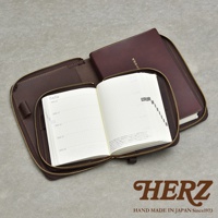 深棕色皮革記事本收納包，拉鍊部分打開，露出內頁有格子和文字的記事本，右側為封閉的記事本，下方有HERZ品牌標誌。