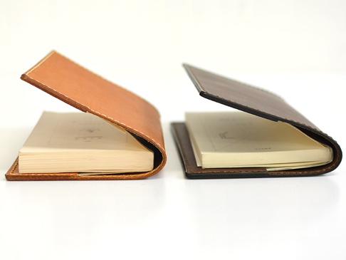 左為淺棕色皮革書套，右為深棕色皮革書套，兩者皆為厚革製成，搭配白色內頁書籍，展示側邊與書脊部分。