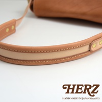 一條 tan 色皮革郵差包的肩帶，帶有雙色拼接和縫線細節，以及兩個黃銅扣環。