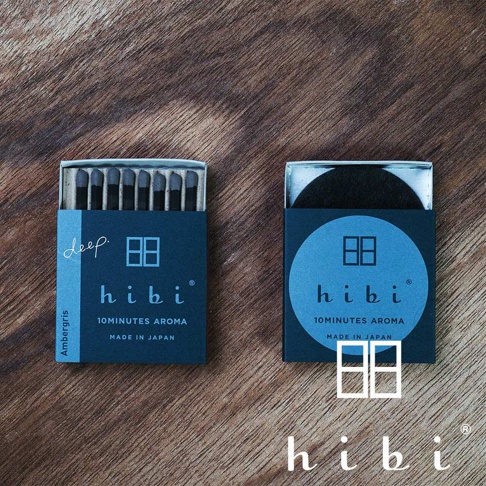 兩個 hibi 品牌香氛火柴盒平放在木質紋理桌面上，左側包裝盒內整齊排列著深色火柴，側面標示著 Ambergris 與 deep 字樣；右側包裝盒則放置了一個黑色圓形燃燒墊，下方以大字體印有品牌標誌與 10 分鐘香氛的說明，整體呈現日式極簡風格。