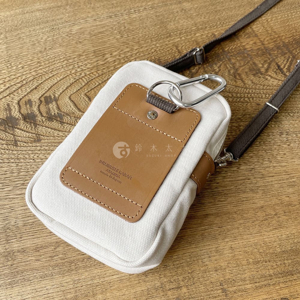 石洗帆布斜背手機包，主體為米白色帆布材質，搭配咖啡色皮革識別證套與肩帶，識別證套上印有品牌標誌與文字，並以金屬掛環固定。