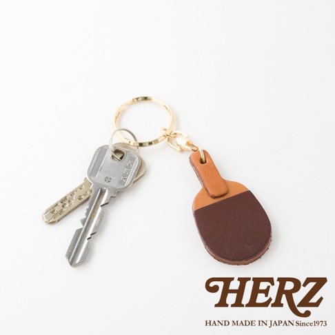 職人手作皮革桌球拍造型鑰匙圈，採用棕色與淺棕色皮革拼接設計，搭配金色鑰匙圈與扣環，掛有兩把銀色金屬鑰匙。