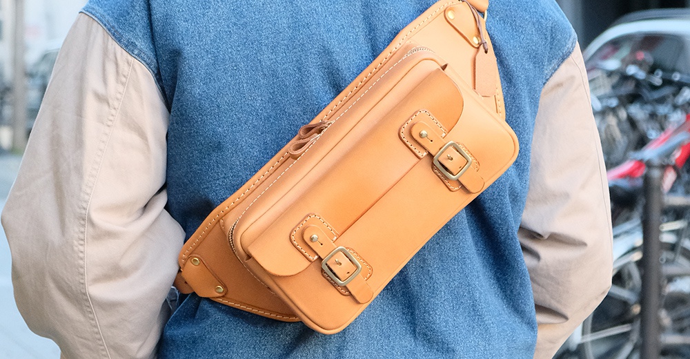 職人手作的淺棕色皮革斜背包，具有雙搭扣設計，以橫跨身軀的方式佩戴。