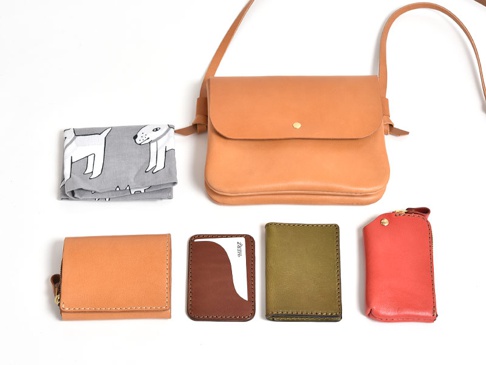 職人手作皮革直式隨行袋 (CP-9) 搭配了多個皮革配件，包括一個淺棕色隨行袋、一個淺棕色摺疊皮夾、一個棕色卡夾、一個橄欖綠卡夾，以及一個珊瑚紅鑰匙包。另有一塊灰色布料，印有白色狗狗圖案。