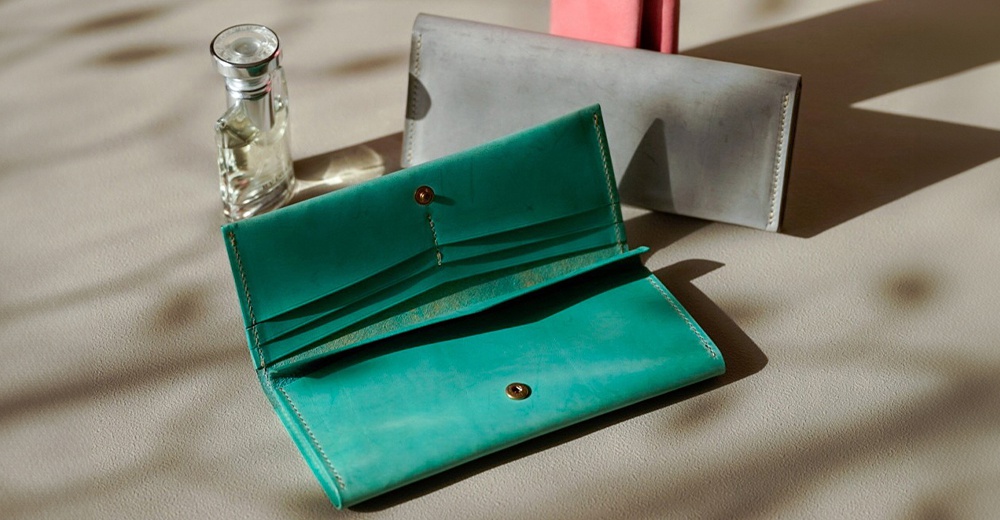 北海道KASANE職人手作義大利Sibilla皮革長夾，展現綠松石色和淺灰色，瓶裝香水放置其中，陽光投下光影，突顯精緻皮革質感。