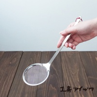 一只手正持著一個銀色不鏽鋼萬用撈油勺，勺頭為橢圓形，內嵌細密網篩，勺柄末端有一個紅色掛環。