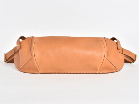 簡約的淺棕色皮革斜背包，包身呈圓潤的長條狀，側面有皮革拼接與白色縫線裝飾，搭配可調節的皮革背帶與金屬扣環。