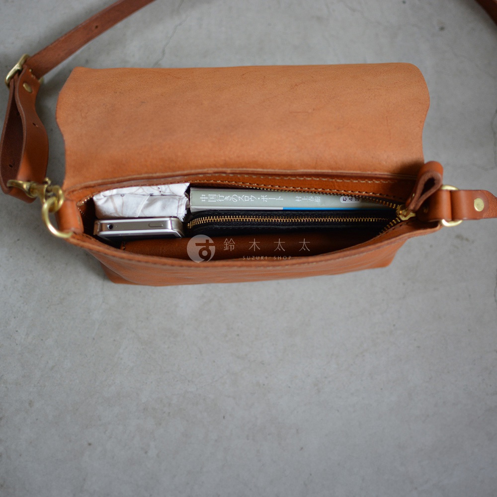 一個棕褐色皮革製的斜背包，包包開口朝上，裡面整齊地擺放著手機、一本書和一些物品。