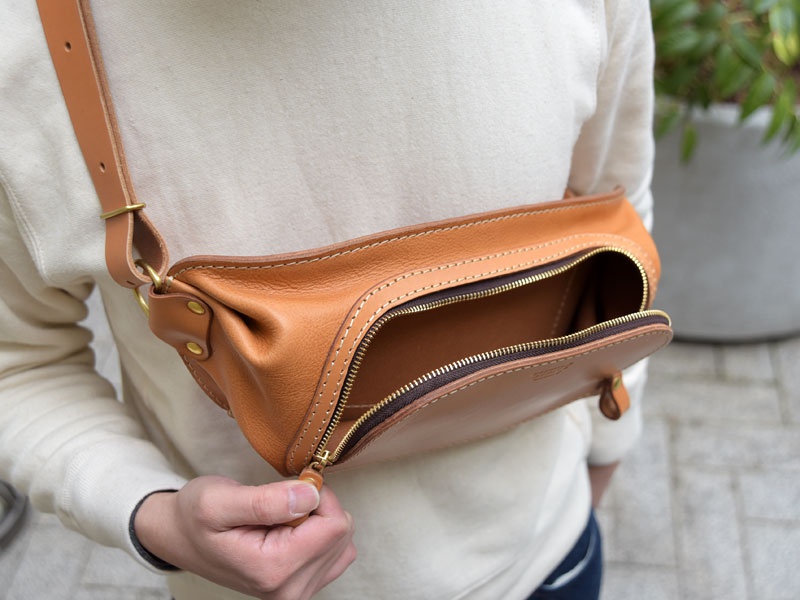淺棕色皮革斜背包，配有黃銅拉鍊和帶有黃銅扣環的皮革肩帶，展示其寬敞的開口。