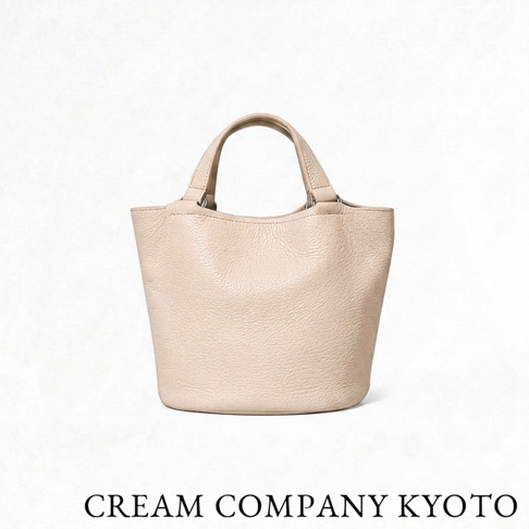 正面視角拍攝的 CREAM COMPANY KYOTO 米白色牛皮革手提水桶包，包身表面呈現細膩的荔枝紋理，採用極簡圓弧水桶造型搭配固定式提把，下方印有品牌名稱。