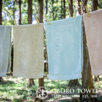 四條懸掛在繩索上的毛巾，顏色由左至右分別為淺灰綠、米黃、嬰兒藍和淺褐。毛巾材質看起來柔軟蓬鬆，底部有細緻的橫條紋裝飾。背景為模糊的樹林。