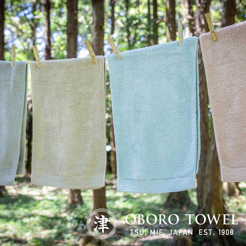 四條懸掛在繩索上的毛巾，顏色由左至右分別為淺灰綠、米黃、嬰兒藍和淺褐。毛巾材質看起來柔軟蓬鬆，底部有細緻的橫條紋裝飾。背景為模糊的樹林。