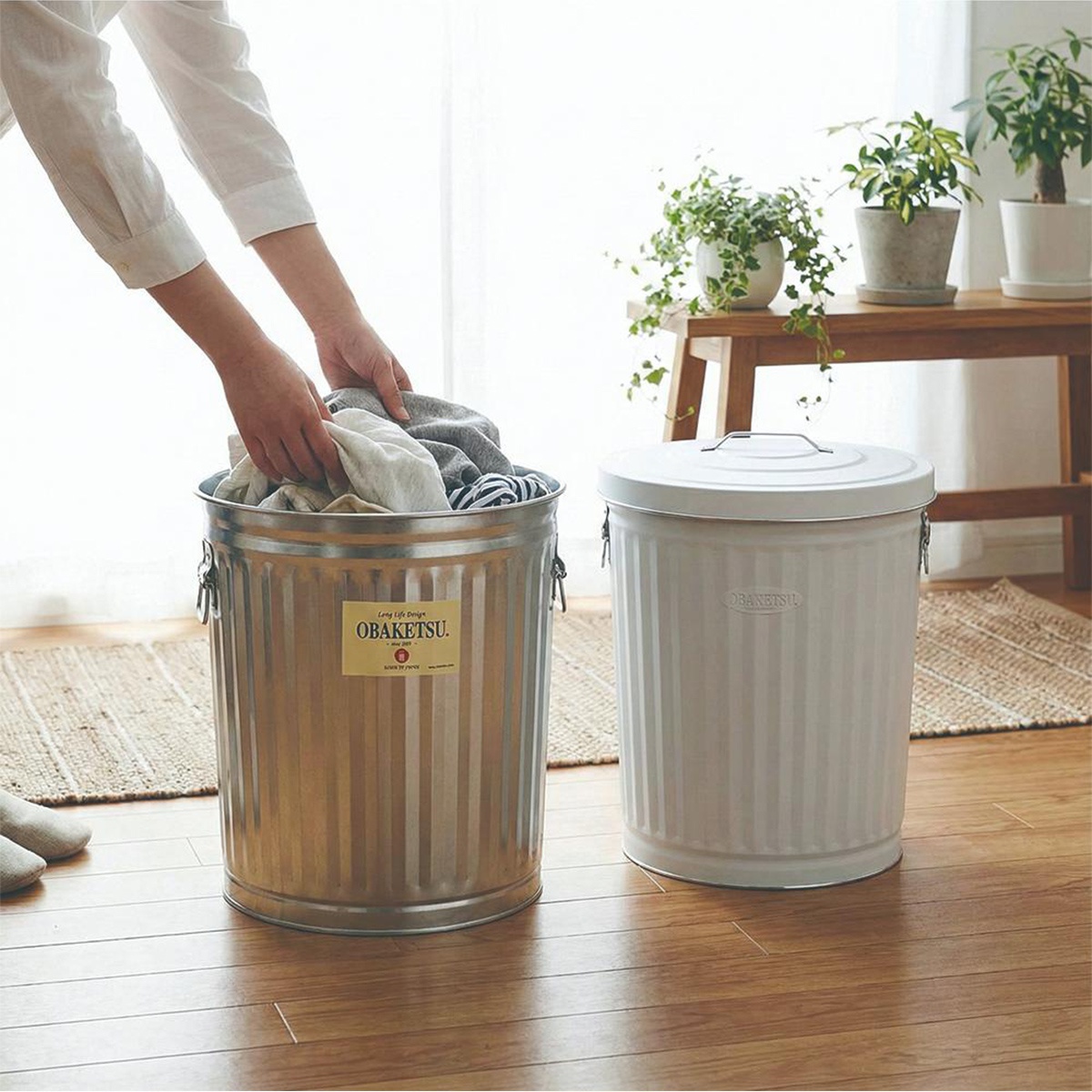 一隻手正將衣物放入左側的金屬腳踏車造型垃圾桶，右側則放置一個白色的同款垃圾桶。