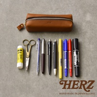 職人手作的棕色皮革Organ箱型收納筆袋，搭配銀色拉鍊，袋上印有「Organ」字樣。筆袋前方擺放著一管黃色UHU膠水、一把金色剪刀，以及一系列不同顏色和款式的筆，包括藍色、銀色、黑色、黃色、藍色、紅色和一支黑色簽字筆。