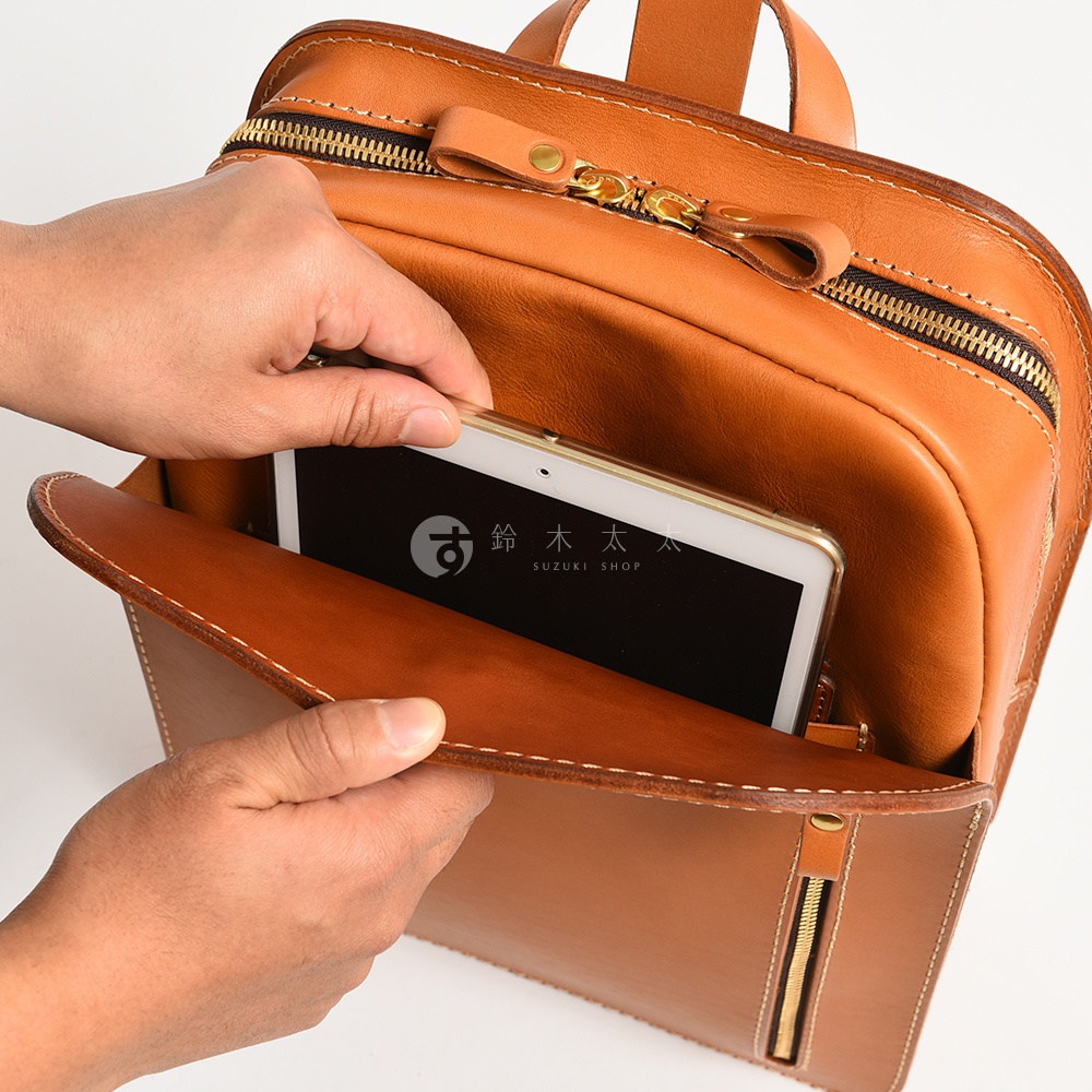 棕色皮革後背包，有金色拉鍊及縫線細節。一隻手正在將平板電腦放入背包前袋。